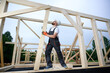 © Василь Івасюк - Construction worker installing wooden beam at house frame construction site. Carpenter in safety gear building timber structure for new home outdoors.