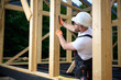 © Василь Івасюк - Construction worker checking wooden beam alignment at timber frame house site. Builder in safety gear holding helmet and using tools while working on wooden structure outdoors.