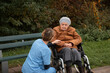 © Anetta - Elderly woman on wheelchair talking with a happy smile to caretaker in blue uniform while taking a break from walk in the park