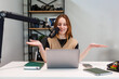 © True Pixel Art - A young woman records at a white desk, speaking into a boom mic, hands raised, with laptop, headphones, and notes, shelves of cameras and books, soft even lighting.