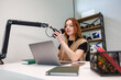 © True Pixel Art - A young woman talks into a cardioid mic on a boom arm at a white desk in a compact studio. A laptop, notebook, and phone sit nearby as soft light gives shallow depth.