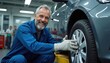 © Pete - Smiling auto mechanic works on car tire in repair shop. Experienced technician wears blue uniform and gloves, checks wheel nuts at garage. Another workers in background help fix vehicles.