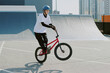 © AnnaStills - Teenage Caucasian boy riding BMX bike performing wheelie trick on urban skatepark ramp, wearing helmet and casual clothing, focused expression, outdoor city background