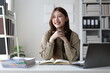 © Wasan - Young Asian businesswoman holding a pen and smiling while thinking about new ideas, sitting at her desk in office with a laptop and documents.