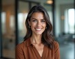 © Maryna - Smiling woman with shoulder-length brown hair in modern office. Soft light highlights features. Wears sleek casual outfit. Background features glass walls, blurred workspace, conveying