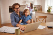 © Stockphotodirectors - A dad sits at a wooden table with his child on his lap, working on a laptop. In the background, a woman prepares something in the kitchen.