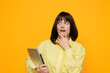 © SHOTPRIME STUDIO - A thoughtful woman stands against a bright orange backdrop, wearing a pale yellow shirt and holding a tablet. She gazes upward with a curious expression, suggesting planning, discovery, or a moment