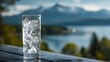 © sungedi - Tall glass filled with clear, cold liquid and ice cubes rests on a weathered wooden surface overlooking a scenic mountain lake.