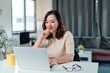 © amnaj - Asian woman concentrating on work while typing on laptop keyboard at home office desk, embracing freelance productivity