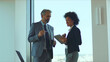 © Videophilia - Two business professionals, diverse man and woman, standing in a bright office reviewing a tablet together, demonstrating teamwork, modern communication and collaborative planning