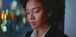 © Videophilia - Young mixed-race businesswoman with curly hair standing near a window, reflecting on her work or career, with blurred city lights creating a bokeh effect in the background