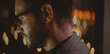 © Videophilia - Man with beard and glasses concentrating on a task, illuminated by a screen, with blurred city lights creating bokeh in the background, symbolizing dedication and working extended hours