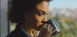 © Videophilia - Thoughtful woman sipping morning coffee by a window, gazing at the cityscape and savoring a quiet, reflective moment before starting the workday