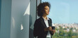 © Videophilia - Confident african american businesswoman in a black blazer pauses with coffee by an office window, gazing at the city skyline while planning strategy and future success