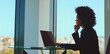 © Videophilia - Thoughtful businesswoman looking out of a large office window, contemplating new ideas and future strategies while working on a laptop, with a cityscape providing a background view