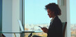 © Videophilia - Black businesswoman sitting by an office window, smiling while using smartphone with laptop on glass table and city view, conveying modern professional confidence and connectivity