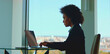 © Videophilia - Young black businesswoman attentively typing on a laptop, dedicating focus to her work from an executive office with a panoramic city view from a large window