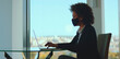 © Videophilia - Young black businesswoman wearing protective face mask diligently working on laptop computer during coronavirus pandemic while sitting at desk in modern office with city view background