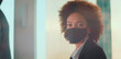 © Videophilia - Professional black businesswoman with afro hair wearing a protective face mask while working in a modern corporate office environment during a global health crisis