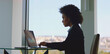 © Videophilia - Young businesswoman with afro hair focused on laptop, analyzing financial data from a high-rise office with panoramic city view, embodying productivity and professional ambition