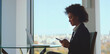 © Videophilia - Young professional businesswoman sitting at a glass table near a large window, using a smartphone and smiling, with a laptop displaying financial data beside her and a city view in the background