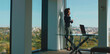 © Videophilia - Confident businesswoman talking on a mobile phone while standing by a large office window, looking out over a sprawling city skyline and green park on a sunny day