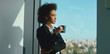 © Videophilia - Young black businesswoman standing near a large office window, holding a coffee mug, looking thoughtfully at the cityscape below while taking a moment during a busy workday