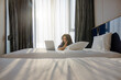 © shine - Smiling asian middle age woman is focused on her laptop screen at  Light cozy bedroom, bed with white sheets and pillows. The sun shines through the window and falls into the bedroom. Comfortable cozy