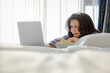 © shine - Smiling middle age asian woman using laptop in bedroom, browsing internet, shopping, chatting online in social network, with popcorn, sitting near window in the early morning in the rays of sun.