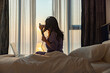 © shine - Beautiful middle aged woman styles her long, healthy hair at home or hotel with a modern hair curler, sitting comfortably in bed and working on laptop. Beautiful Sunrise through the curtains on window