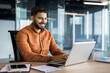 © Liubomir - Young indian businessman smiling and typing on a laptop, sitting at a desk in a contemporary office reflecting productivity, success, and professional communication
