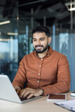 Indian businessman working on a laptop, standing at a desk in a contemporary office setting with glass walls, reflecting a positive attitude and professional success while engaging in business tasks