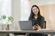 © PRIME STOCK LAB - Young woman smiling while working at laptop and holding smartphone in bright home office with plant and notebook