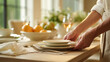 © Lisa - Close-up of a person arranging white ceramic plates and linen napkins on a wooden dining table with fruits and flowers in soft natural light, creating an elegant and warm table setting