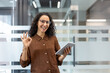 © Liubomir - Businesswoman with curly hair and glasses holding a digital tablet, smiling and giving an optimistic okay gesture, symbolizing success and approval in a modern office environment