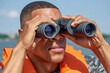 © azwan - Serious young African American man in orange life vest looking through binoculars outdoors