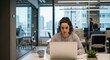 © NanieStudio - Focused young businesswoman with headphones working on laptop in modern open space office. Concentrated female professional listening to webinar, e-learning, or on a video call.