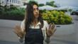 © Krakenimages.com - Woman with palms raised and hands visible wearing denim apron on a street near planters and umbrellas, palms facing camera; concern.