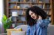 © Liubomir - Young woman sitting on a sofa in a living room massaging a stiff neck with her hand, showing discomfort and stress from poor posture and sedentary home life