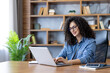 © Liubomir - Smiling young woman with curly hair and glasses, sitting at a desk and typing on a laptop, enjoying remote work from a comfortable home office environment