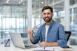 © Liubomir - Smiling bearded man in a casual blue shirt and white t-shirt sitting at a contemporary office desk, holding his eyeglasses, and looking directly at the camera with a laptop and tablet nearby