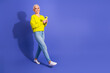 © deagreez - Bright senior woman in yellow sweater walks and smiles while using smartphone against purple background