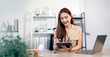 © NAMPIX - Asian businesswoman smiles while using a stylus on a digital tablet, with a laptop nearby on the office desk.