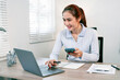 © NAMPIX - Young Asian businesswoman smiles while working on her laptop and phone in a modern office.