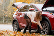 © MarijaBazarova - Woman checking flat tire and inspecting wheel of red SUV car during autumn roadside breakdown emergency on country road