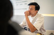 © Daniel - Asian man in a white shirt holding a notebook and pen while thinking deeply at a modern café table, with a laptop in front of him, showing a focused and contemplative expression in a bright setting
