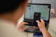 © Daniel - Asian man using a smartphone while sitting in front of a laptop with a messaging app on the screen, multitasking between phone and computer in a modern workspace with blurred background