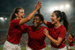 © OlenaSpark - Three female soccer players (european, african-european, asian) celebrating goal with joy and hugs on a brightly lit evening stadium, red jerseys and white shorts, emotional faces