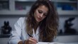 © Thares2020 - Young female professional wearing a lab coat focused on diligently writing notes in a scientific laboratory notebook at her desk