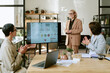 © AnnaStills - Caucasian middle aged woman standing and presenting business analytics on large screen to two Caucasian middle aged women, sitting at table and clapping during office meeting